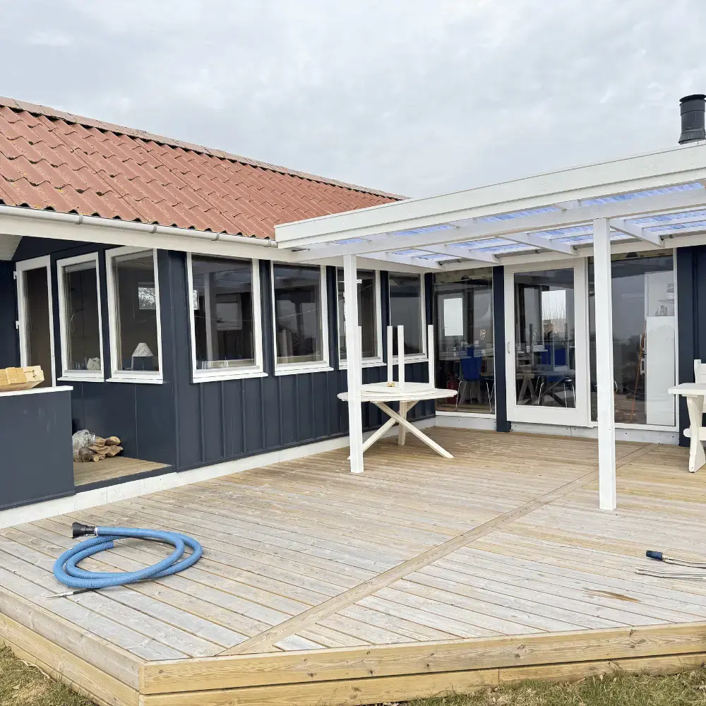 Rent rødt tegltag på et mørkeblåt sommerhus med hvid pergola over træterrassen efter tagrens