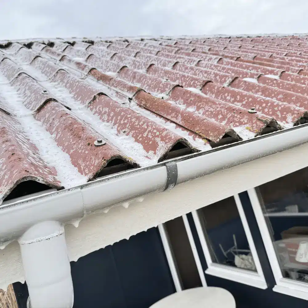 Rødt betontegltag dækket af hvidt rensemiddel set fra tagfoden, med hvid tagrende og nedløb på et mørkeblåt hus