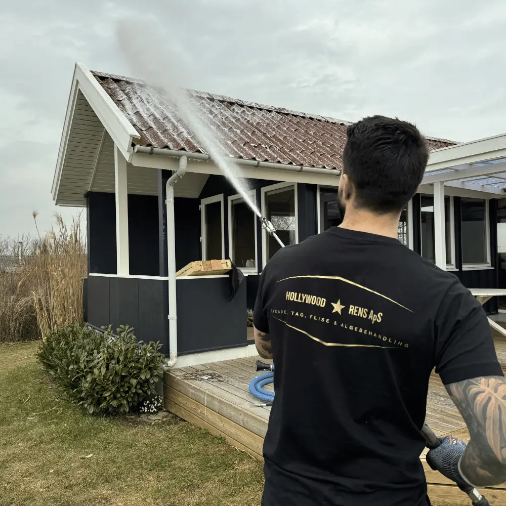 Medarbejder i sort Hollywood-trøje sprøjter taget på et mørkt sommerhus med hvide gavle og træterrasse