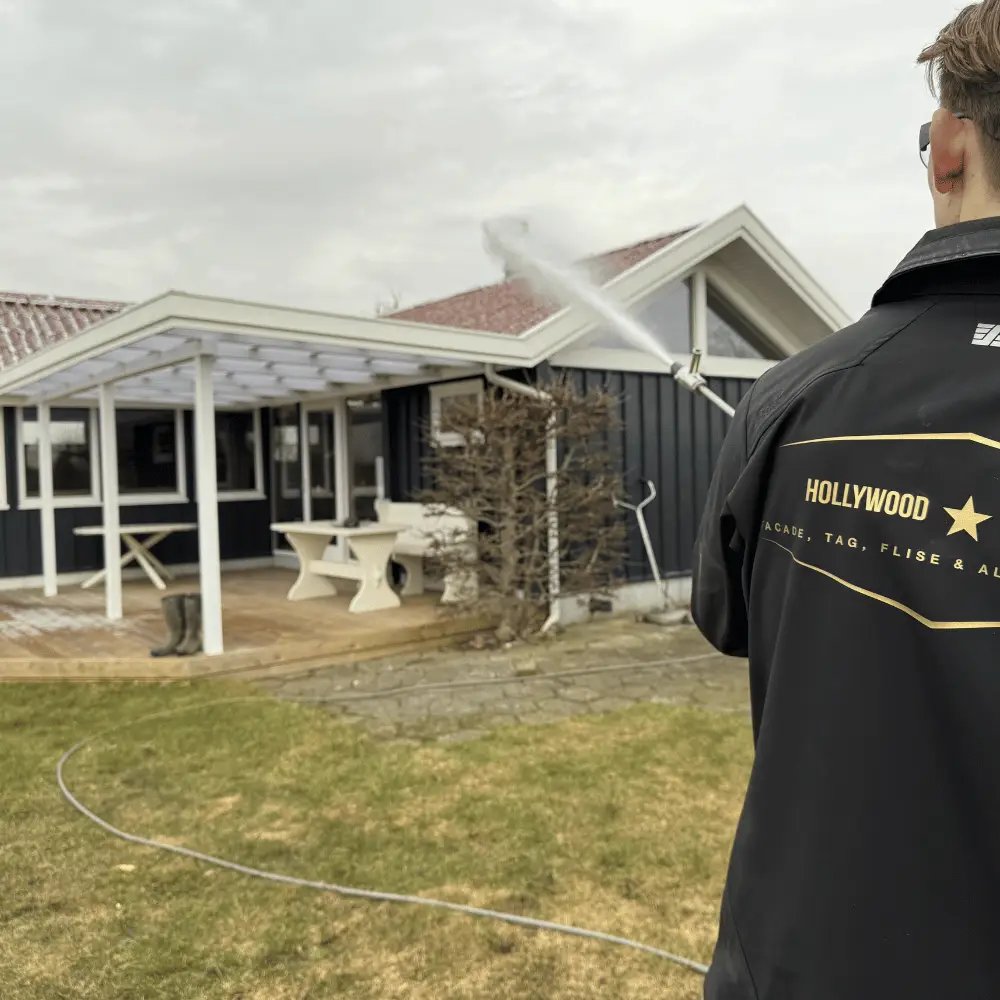 Medarbejder i Hollywood-jakke sprøjter med teleskoplanse et rødt tegltag på et mørkt hus med hvid pergola